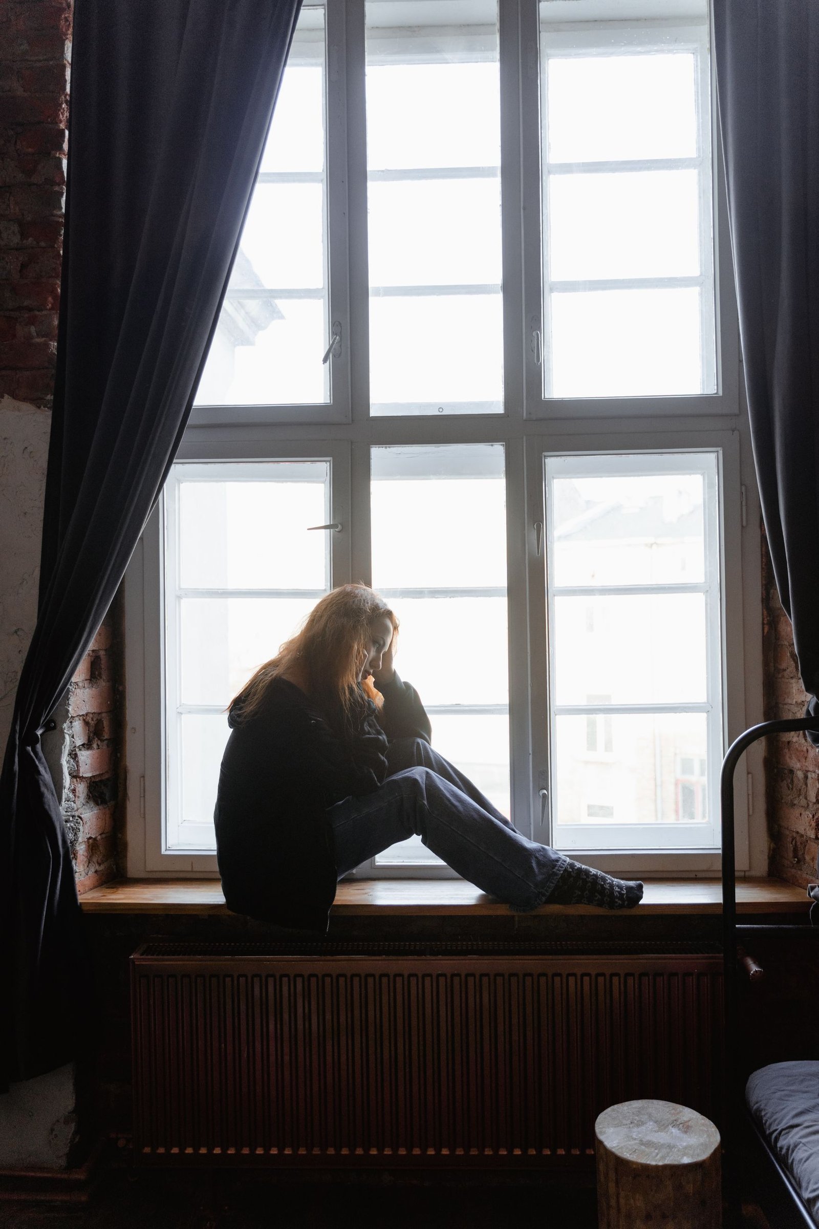 A woman sits thoughtfully on a window sill, gazing outside with curtains open.