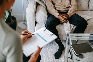 Blurred unrecognizable female therapist taking notes on clipboard while sitting in front of ethnic male patient with clasped hands during meeting